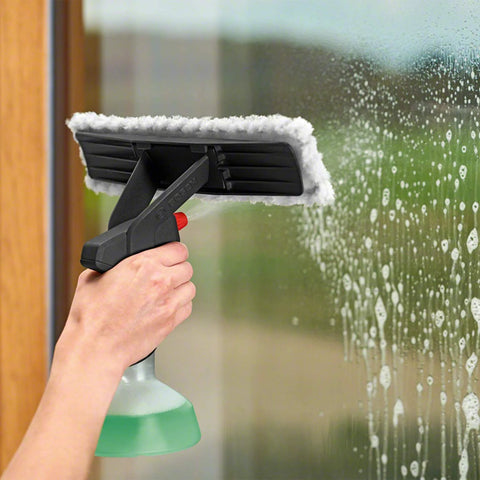 GlassVAC window cleaning tool with black pad and white microfiber edge, hand-held over soapy glass with a green spray bottle.