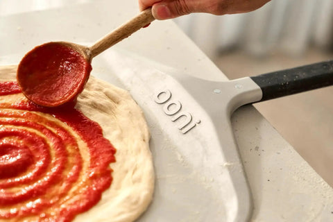 Compact 12 Inch Aluminum Pizza Peel With Handy Handle For Easy Pizza Turning And Oven Prep Lightweight Yet Durable Ideal For Home Kitchens And Ooni Ovens Space Saving Design For Quick Pies in use spreading sauce on dough.