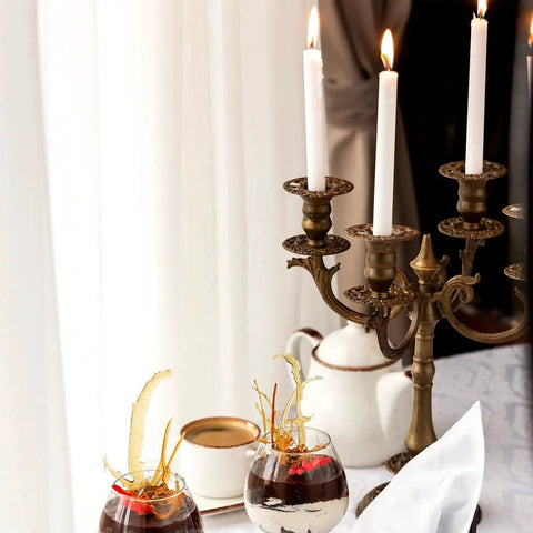 An elegant table setting with a brass candelabra, white candles, dessert glasses, and a teapot on a white tablecloth.