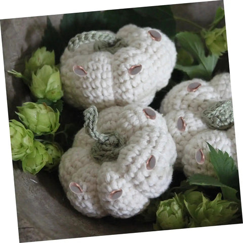 Crocheted white pumpkins with green stems and embellishments surrounded by green foliage, displayed in a decorative setting.