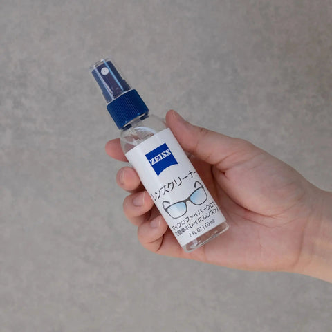 Hand holding ZEISS Lens Cleaner 2oz Spray bottle for coated lenses and optics.