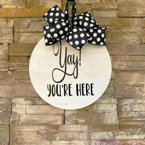 Round wooden sign with 'Yay! You're Here' text, decorated with a black and white polka dot bow, mounted on a brick wall.