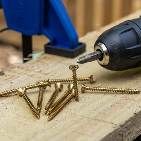 Close-up of M4 X 50mm Wood Screws Flat Head Phillips Self Tapping Carbon Steel Zinc Plated next to a drill on wood surface