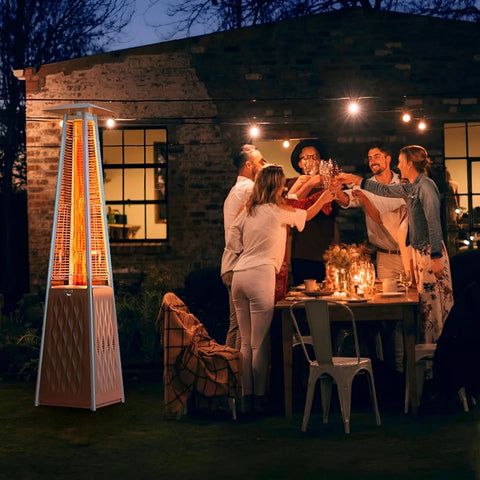 Patio propane heater tower, beige metal with glowing flame, left of a nighttime backyard dinner party.