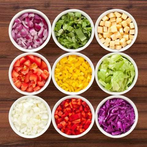 Nine bowls of chopped vegetables and cheese on a wooden surface, showcasing preparation with the all in one vegetable chopper and prep station