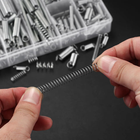 Extension spring, zinc-plated steel, held by fingers; background shows a plastic tray of assorted springs.