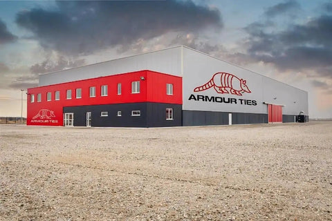 Exterior view of a large red and gray industrial building with 'Armour Ties' logo, situated in a gravel lot under a cloudy sky.