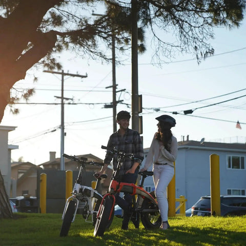 Two electric bikes with riders in a park; white and red bikes with helmets.