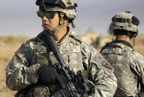 Two soldiers in camouflage uniforms with tactical gear and helmets, one holding a rifle in a desert setting.