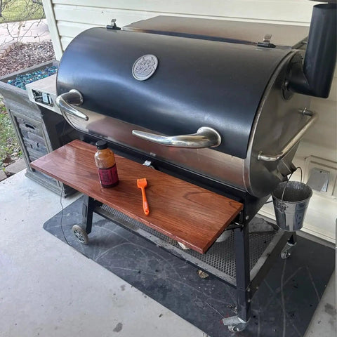 Black outdoor BBQ grill with attached wooden shelf, featuring grilling tools and spice jar on the porch setting.