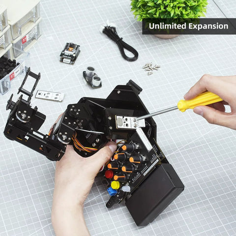 Robotic arm kit with Arduino controller being assembled; black multi-joint arm, wires, yellow screwdriver on gray grid mat.
