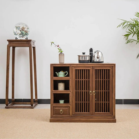 Wooden cabinet with slatted doors and shelves, metal handles, topped with teapots, next to a plant and pedestal with vase