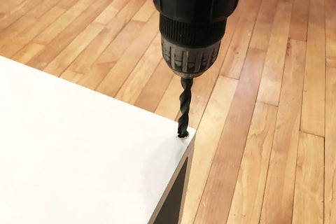 Drill creating hole in white wood surface on hardwood floor, preparing for Brown Wood Wheel Stem Casters Set of Two installation.