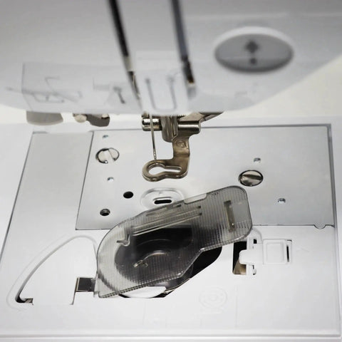 Bobbin cover plate area on a sewing machine; front view shows exposed bobbin housing, metal needle plate, and clear presser foot.