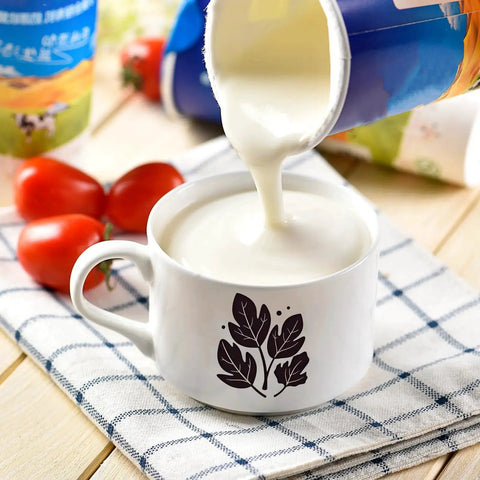 White mug with black leaf design filled with creamy yogurt pouring from a container, tomatoes in the background, on a checkered cloth.