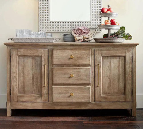 Wooden sideboard with drawers and cabinets featuring brass knobs and handles, decorated with glassware and a fruit display.