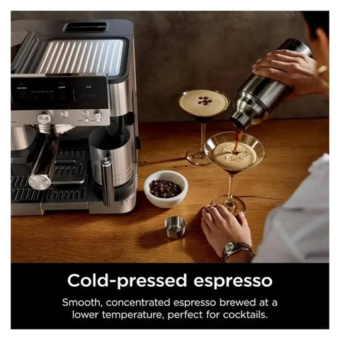 Person pouring cold-pressed espresso into glass beside coffee machine with beans on wooden table