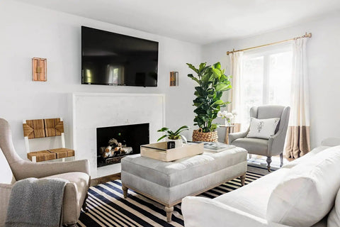 Modern living room with a wall-mounted TV above a fireplace, surrounded by grey and white furniture, houseplants, and natural light.