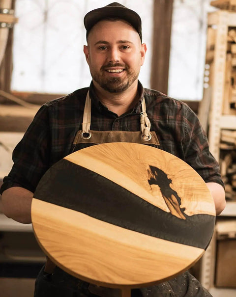 Woodworker holding round wooden table with black resin inlay in workshop, showing craftsmanship and design.