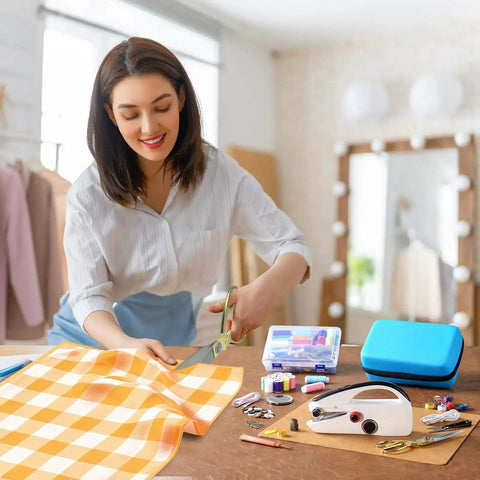 Woman using Heavy Duty Handheld Sewing Machine Kit With Blue Leather Storage Case And 110 Piece Sewing Kit, fabrics and sewing tools displayed.