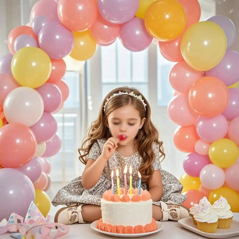 Colorful balloon arch with a child sitting in front, cake with candles, and cupcakes beneath the arch indoors.