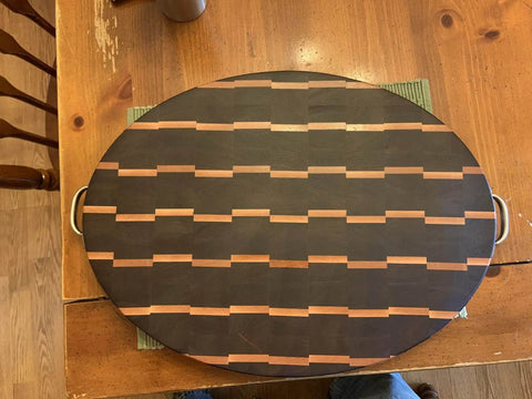 Wooden oval cutting board with dark and light wood pattern, handles on sides, displayed on wooden table.