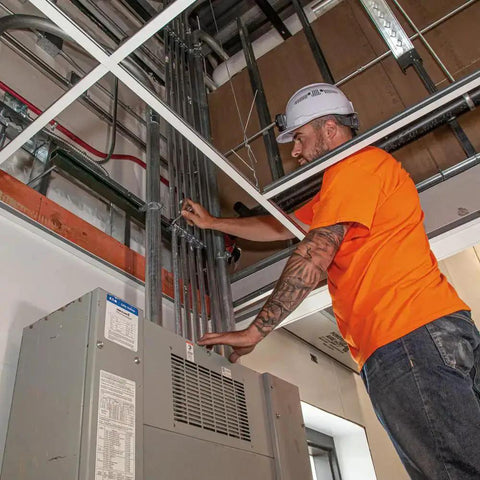 Construction worker in orange shirt adjusting wiring in a building ceiling.
