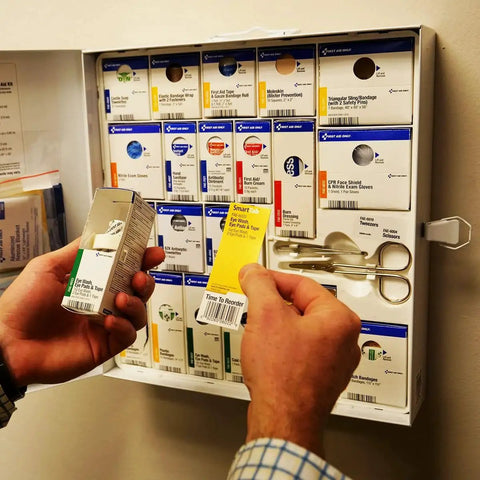 Hands holding 4 Piece Eye Wash Kit With Oval Eye Pads, Tape And Emergency Eye Rinse in front of a first aid cabinet