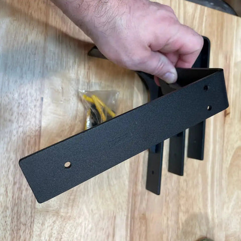Hand holding Heavy Duty 10x6 Inch Shelf Brackets Set Of 4 Sturdy L Shaped Metal Supports, matte black, with screws in background.