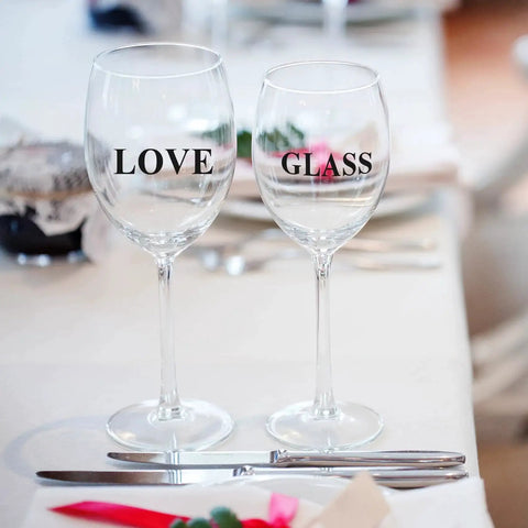 Two wine glasses on a dining table, one labeled 'LOVE' and the other 'GLASS', with silverware and a white tablecloth.