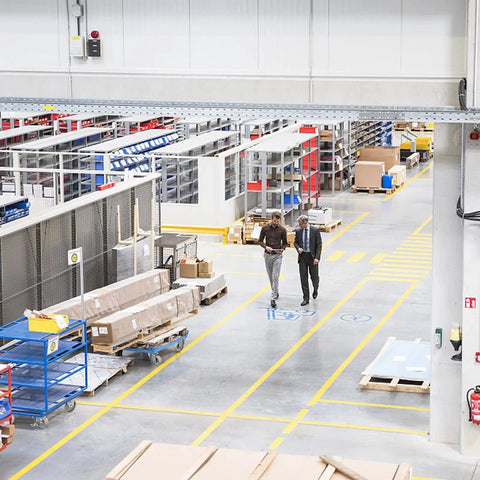 Industrial warehouse with two people walking among shelves, carts, and boxes