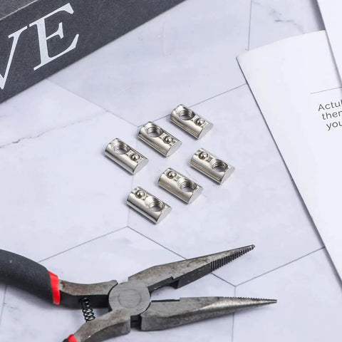 Close-up of 6 roll-in T-nuts with ball spring for 10 series aluminum extrusion profiles, 1/4-20 size, zinc plated steel on table with pliers