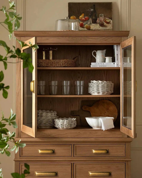 Open wooden kitchen cabinet with glassware, plates, and cutting board; antique brass cup pulls visible on drawers below.