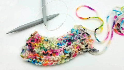 Colorful knitted piece with circular knitting needles on white background, showing multicolored yarn and partial knit project.