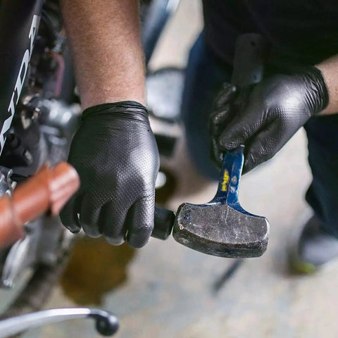 Person wearing Heavy Duty Black Nitrile Gloves 8 Mil Diamond Texture Powder Free Food Safe 1000 using a hammer in a workshop.