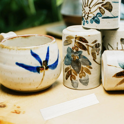 Ceramic pottery mugs and jars, several, with blue floral glaze on a wooden table; white ribbed strip in foreground.