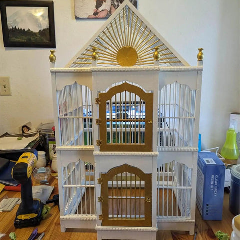 White and gold birdcage with sunburst design and arched doors on a wooden table surrounded by tools and objects.