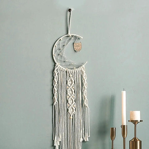 Macrame wall hanging in crescent moon shape with owl charm, featuring intricate knots and tassels, hung on a light blue wall.
