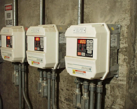 Three industrial electrical panels mounted on a concrete wall, featuring digital displays and high voltage warning signs.
