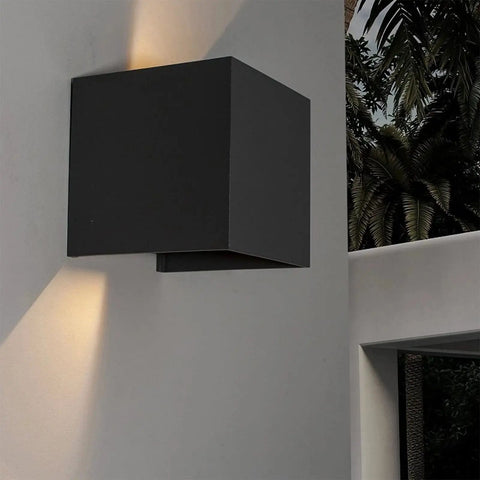 Modern black cube wall sconce emitting light up and down, mounted on a white wall, outdoor setting with trees in the background.