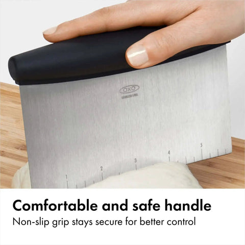 Image of a hand holding a stainless steel all purpose scraper and dough cutter for pizza bread and cookie dough, vegetable chopper with built in quarter inch markings, dishwasher safe kitchen essential for precise prep.