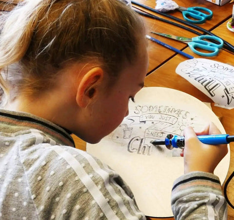 Child drawing on a craft paper circle with markers, surrounding tools visible. 4 Pack 18 Inch Round Plywood Boards for Crafts and DIY Projects.