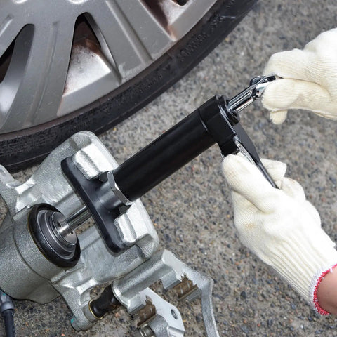 Side view of a pneumatic brake caliper piston tool in use on a car disc brake; black barrel, silver rod, gloved hand.