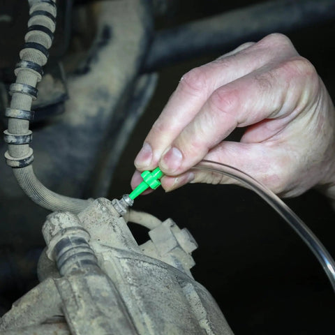 Hand attaching a bright green brake bleeder adapter to a dusty car brake caliper using a clear hose