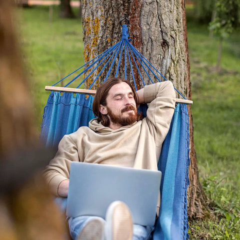 Person relaxing in a blue hammock tied to a tree, holding a laptop, outdoors with Built Tough 5 Inch Eye Hooks Set Of 4 Screws In Carbon Steel Black Large Screw Eye Hooks For Wood And Cables Heavy Duty Gear To Secure Wires Outdoor And Indoor Use With Anti Rust Finish.