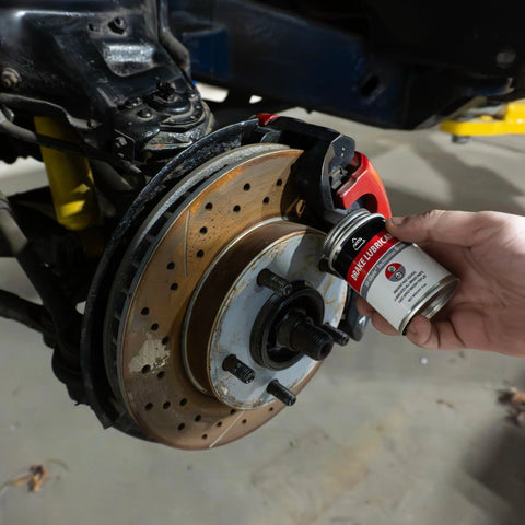 SIL-Glyde 4oz silicone brake grease can held near a red brake caliper on a drilled rotor, wheel hub and studs visible