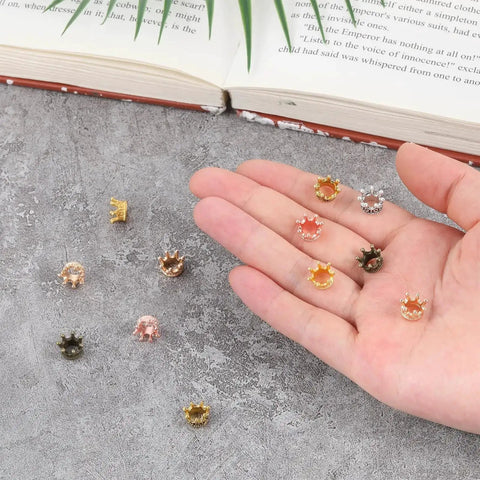 Hand holding 30 Pieces Mini Crown Charms Set For Crafters Mixed Color Vintage 3D Alloy King Crowns Beads on a textured surface.