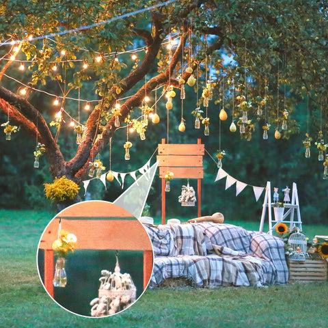 Outdoor garden scene with tree decorated with lights and hanging jars, cozy seating with plaid blanket, wooden crates, and flower arrangements.