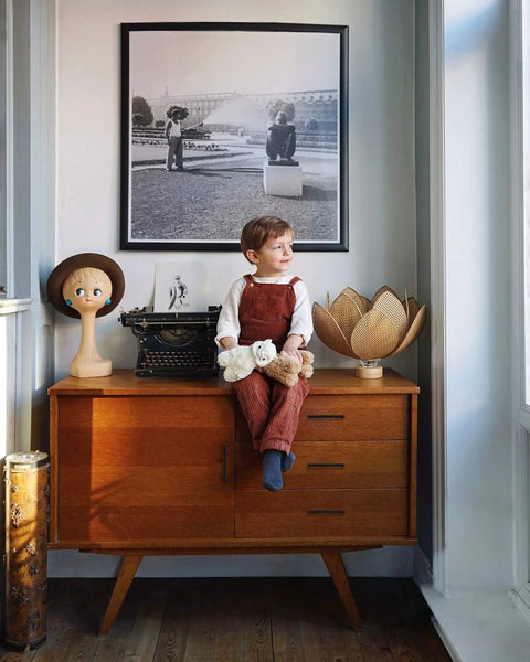Child sitting on a wooden cabinet holding a teddy bear, with decorative items and a framed black-and-white photograph above.