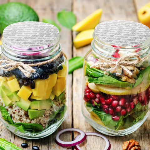 Two glass mason jars with foil-sealed lids and twine on a wooden table, filled with colorful salad ingredients.